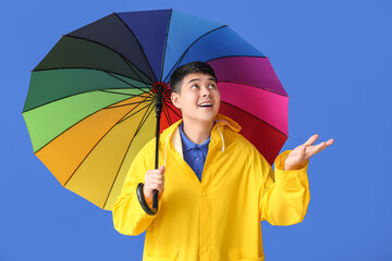 Young Asian man in raincoat and with umbrella on color background