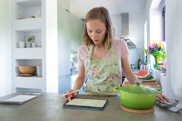 Focused young woman consulting recipe while cooking in her kitchen, using tablet near big saucepan on counter. Front view. Cooking at home and internet concept