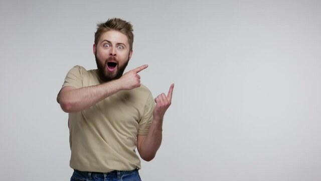 Surprised bearded guy in t-shirt pointing aside and looking with open mouth amazed shocked extremely excited, showing blank copy space for unbelievable advertise. studio shot isolated, gray background