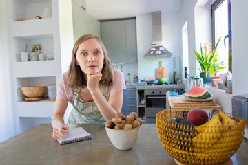 Obraz na płótnie Canvas Pensive woman writing menu in her kitchen, leaning on counter with fruits and nuts and looking at camera. Cooking at home concept