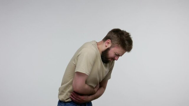 Bearded man feeling painful cramp in stomach, clasping bloated belly and suffering severe abdominal pain, spasm of irritable bowel syndrome of diet. indoor studio shot isolated on gray background