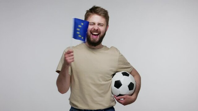Joyful happy football fan bearded guy in t-shirt holding soccer ball and waving European Union flag, excited about winning goal, cheering for favorite sport game. studio shot isolated, gray background