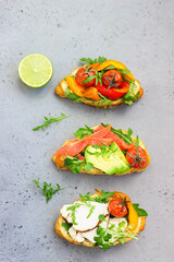 Variety of croissant sandwiches with grilled pepper, tomatoes, smoked salmon, turkey, avocado and arugula served with micro green on grey concrete background. Top view.