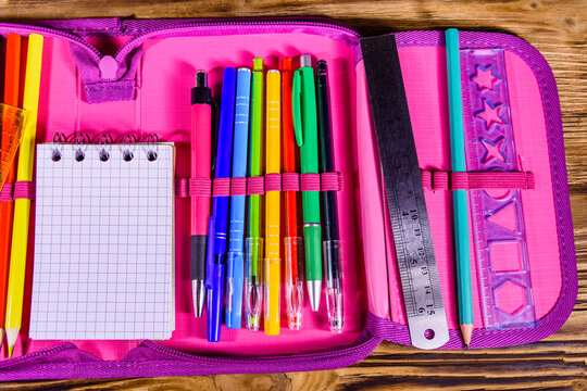 Different school stationeries (pens, pencils, notepad, ruler and protractor) in a pink pencil box. Top view