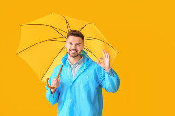 Young man in raincoat and with umbrella showing OK on color background