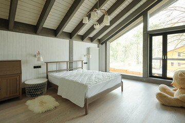 Cozy bedroom with big toy bear, small carpet bedside table and double bed in cottage house with glass wall