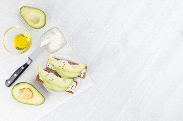 Cooking of fresh light summer appetizer of crisps bread, avocado slices, olive oil, black sesame seeds, cream cheese with ingredients on white wood board, flat lay, copy space.