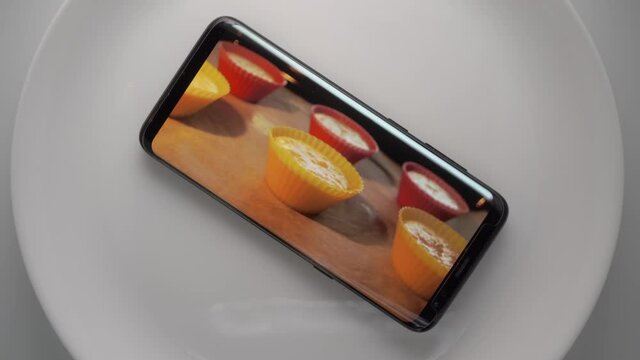 Cell phone on plate with homemade muffins cupcakes on screen. Food blog concept. Top view of mobile phone with food footage on screen rotating.