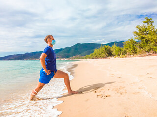 Coronavirus COVID 19. Young beautiful man doing sport performing workouts exercises and squatting near sea in the morning during quarantine. Active life in surgical sterilizing face mask protection