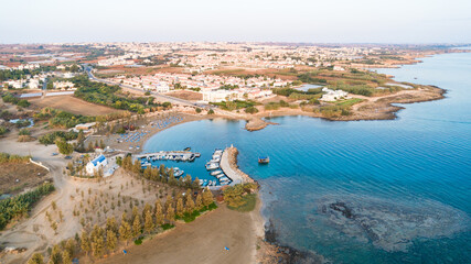 Aerial bird's eye view of coastline sunset, landmark white washed chapel Agia Triada beach, Protaras, Famagusta, Cyprus from above. Ayia Trias bay church and a boat out of the small port at sunrise.