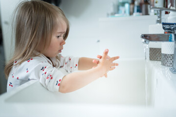 Cute little girl 2-3 years old washing hands with soap and water in bright bathroom.