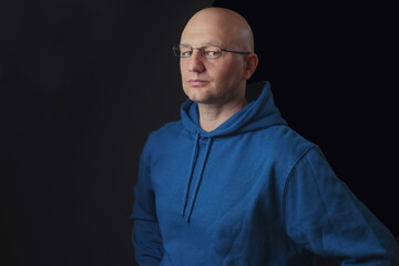 Portrait of bald and clean shaven man with glasses dressed in blue hoodie on a black background.