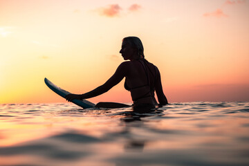 Portrait from the water of surfer girl with beautiful body on surfboard in the ocean at colourful sunset time