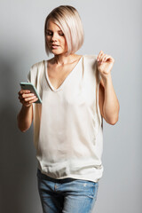 A young blonde girl in jeans and a white T-shirt with a phone in her hand looks at the screen. Blogging, online communication and social networks. Gray background. Vertical.