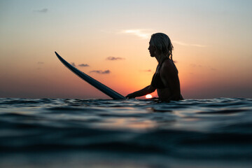 Portrait from the water of surfer girl with beautiful body on surfboard in the ocean at colourful sunset time