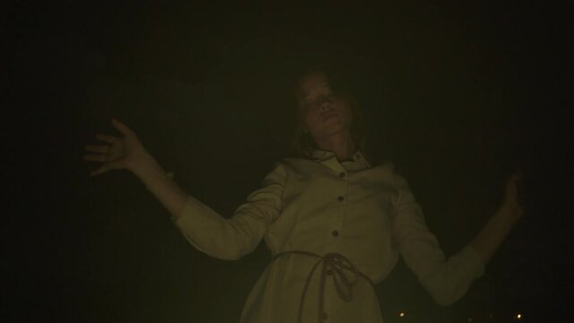 Slow ecstatic dance of young beautiful long haired woman in white dress in the darkness. Yellow light. Low angle shot. Dancing before the fire on city background.