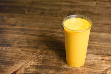 Summer yellow cocktail of mango, banana, ice and juice in a plastic glass on a wooden table with copy space