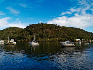Fototapeta na wymiar Beautiful morning view of Creek with boats and reflections of blue sky, light clouds, mountains and trees. Berowra Waters, Berowra Valley National Park, New South Wales, Australia