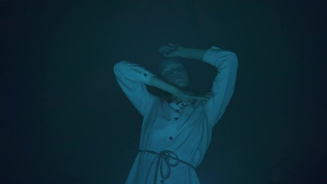 Slow ecstatic dance of young beautiful long haired woman in white dress in the darkness. Blue light. Low angle shot. Night club dancing party. Freedom mood.
