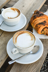 Two cups of cappuccino with latte and croissant on wooden background. stylish toning. morning coffee for couple in love.