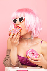 Stylish girl in a pink wig eats delicious donuts. Shot in a studio on a pink background.
