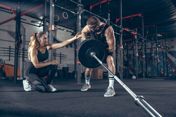 Fototapeta na wymiar Beautiful young sporty couple training, workout in gym together. Caucasian man training with female trainer. Concept of sport, activity, healthy lifestyle, strength and power. Working out with barbell
