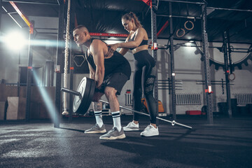 Obraz na płótnie Canvas Beautiful young sporty couple training, workout in gym together. Caucasian man training with female trainer. Concept of sport, activity, healthy lifestyle, strength and power. Working out with barbell
