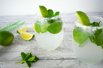 Two glasses of ice lemonade with lime and fresh mint stand on a wooden surface. Near are mint leaves, lime wedges and striped cocktail tubes. The edges of the glasses are covered with salt.