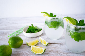 Two glasses with summer cold drinks with lime and mint stand on a light wooden table. Lime slices lie near and there is a white bowl with mint. The edges of the glasses are coated with sugar.