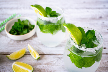 Mojito cocktail glasses with lime and mint stand on a light wooden table. The edges of the glasses are grated with lime and coated with sugar. Lime slices lie near and there is a bowl with mint.