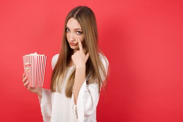 Young caucasian woman holding bucket with popcorn standing over isolated red background Pointing to the eye watching you gesture, suspicious expression.