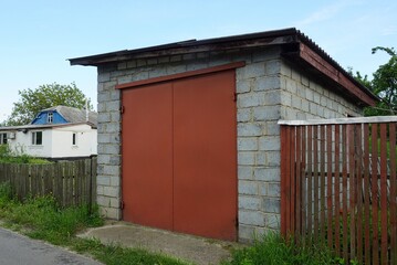 facade of a gray brick garage with red iron closed gates and a wooden fence on a rural street