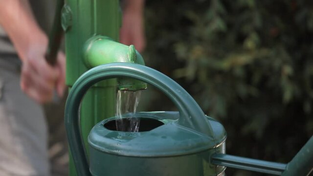A man is using a manual water pump in a green environment and filling a watering can with fresh water 