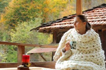 A young woman wrapped in a woollen blanket. She is drinking from a cup. It's a rural scene in a village.