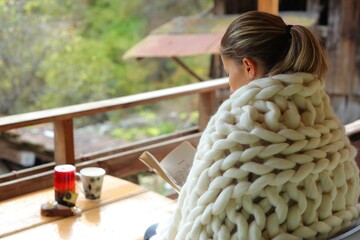 A young woman reading a book. She is cover in a woollen blanket. The photo is taken from behind her shoulder. It's a rural scene in a village.