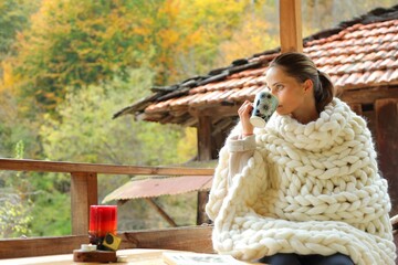 A young woman wrapped in a woollen blanket. She is drinking from a cup. It's a rural scene in a village.