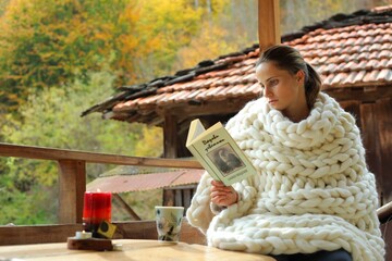 A young woman reading a book. She is cover in a woollen blanket. It's a rural scene in a village.