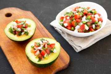 Homemade Pico de Gallo Stuffed Avocado on a rustic wooden board on a black background, low angle view.