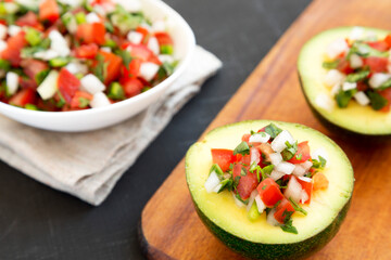 Homemade Pico de Gallo Stuffed Avocado on a rustic wooden board on a black surface, low angle view. Close-up.