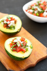 Homemade Pico de Gallo Stuffed Avocado on a rustic wooden board on a black surface, side view.