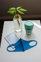 Photo from the top of a reusable face mask, an electronic device a coffee and a plant on a white table