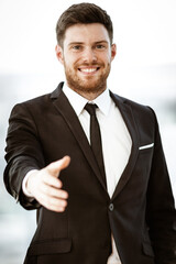 Business concept. Successful young businessman at work. Manager standing in office happy reaches out for a handshake. Man smiling in suit indoors on glass window background