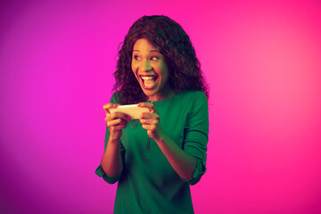 Crazy shocked, holding phone. African-american young woman on gradient pink background in neon light. Beautiful female model. Concept of human emotions, facial expression, sales, ad. Copyspace.