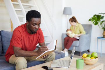 Fototapeta premium African young man reading his notes in note pad while sitting on sofa in front of laptop with woman with baby sitting in the background