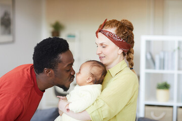 Obraz premium Young woman holding baby on her hands while African man kissing her they are at home