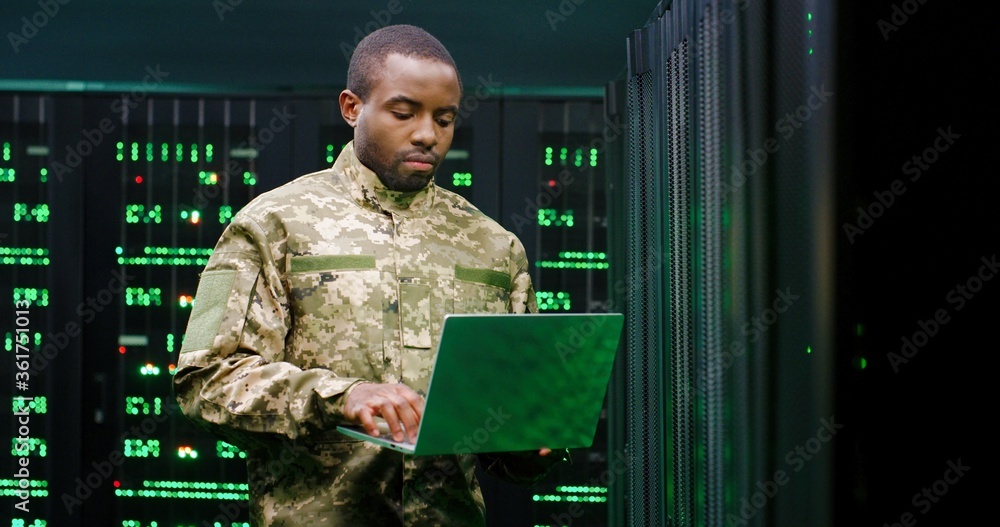 Wall mural us army male african american officer in uniform stading at server wih secret data and using laptop 