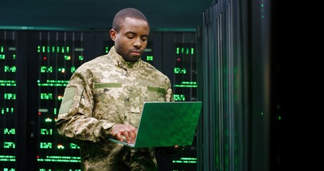 US army male African American officer in uniform stading at server wih secret data and using laptop computer to check the information. Military monitoring service room. Army networking concept.