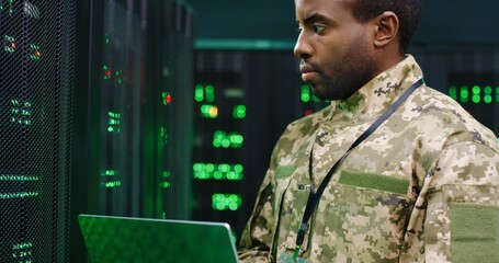 Close up of African American male army technician in camouglage costume and hat typing on keyboard of laptop computer while working with secret data at server room. Government analytic center.