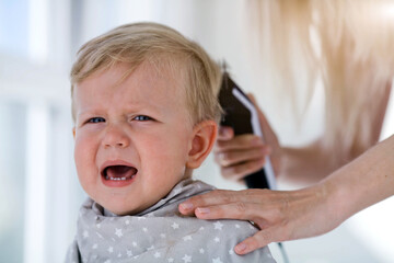 Female hands trim a crying baby with an electric hair clipper in a hairdresser. First haircut.