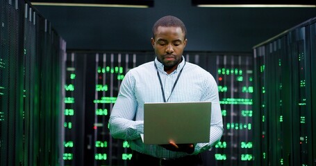 Male system administrator of big data center typing on laptop computer while working in server...